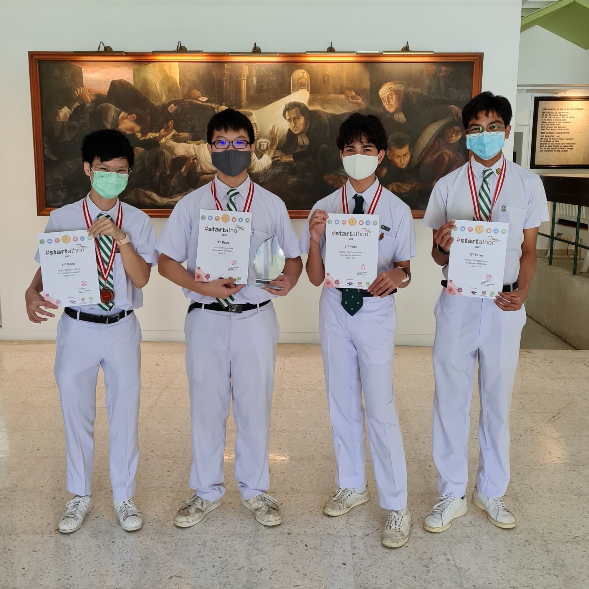 Four masked students in white uniforms hold Startathon 3rd Prize certificates and a trophy indoors.